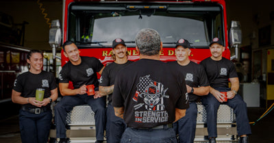 A group of firefighters sitting on the front of a fire truck facing forward. One firefighter standing in the middle facing the fire truck to show the Strength In Service design on the shirt.