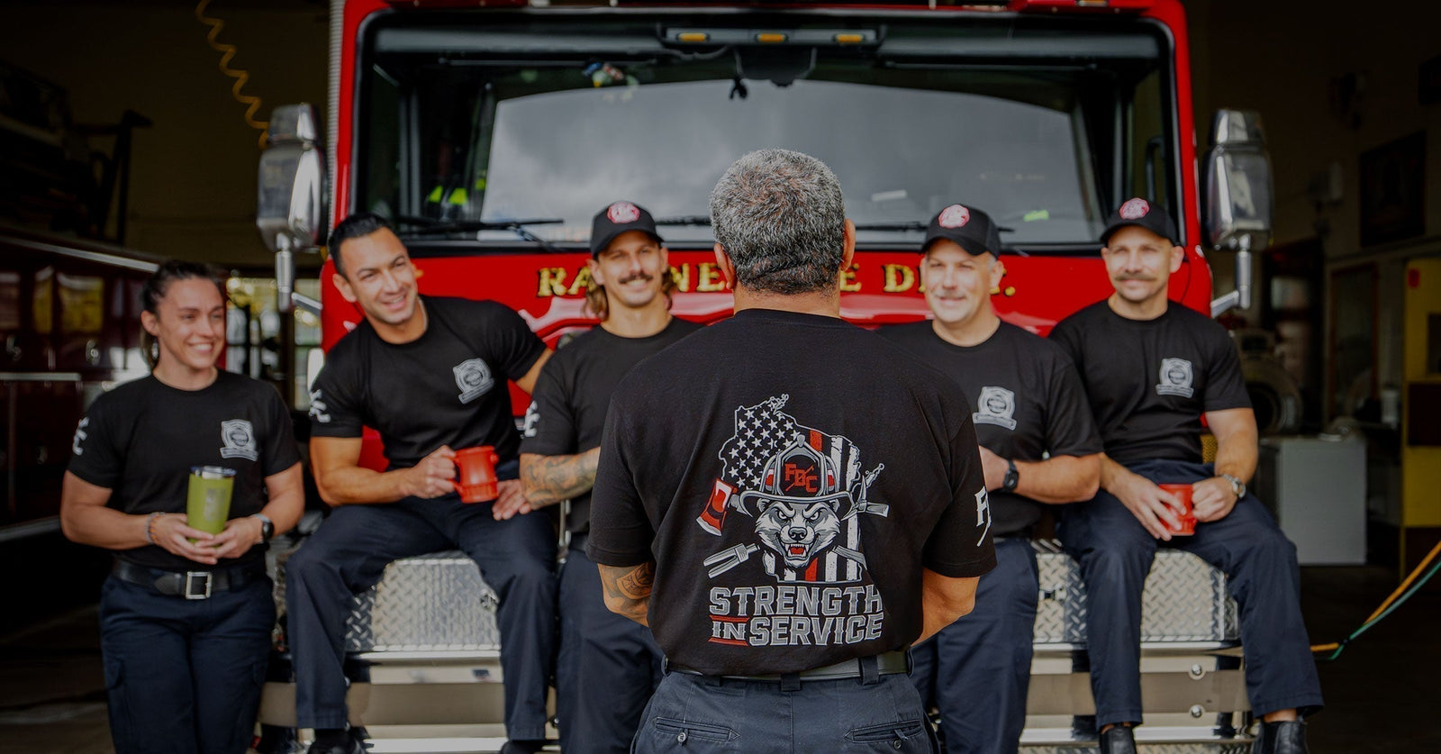 A group of firefighters sitting on the front of a fire truck facing forward. One firefighter standing in the middle facing the fire truck to show the Strength In Service design on the shirt.