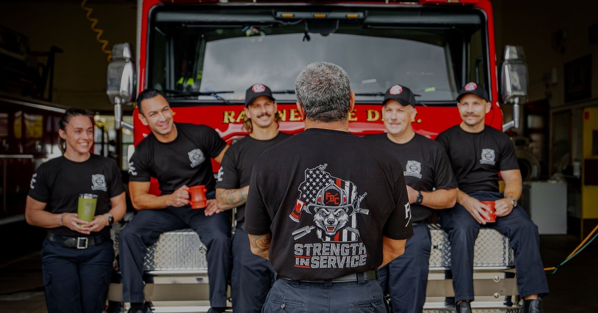 A group of firefighters sitting on the front of a fire truck facing forward. One firefighter standing in the middle facing the fire truck to show the Strength In Service design on the shirt.