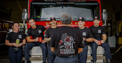 A group of firefighters sitting on the front of a fire truck facing forward. One firefighter standing in the middle facing the fire truck to show the Strength In Service design on the shirt.