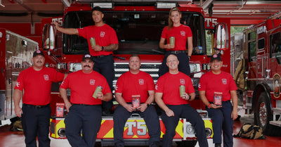 Seven Plano Fire-Rescue firefighters in red t-shirts and navy pants pose with green tumblers and Lone Star Grit coffee bags in front of a fire truck.