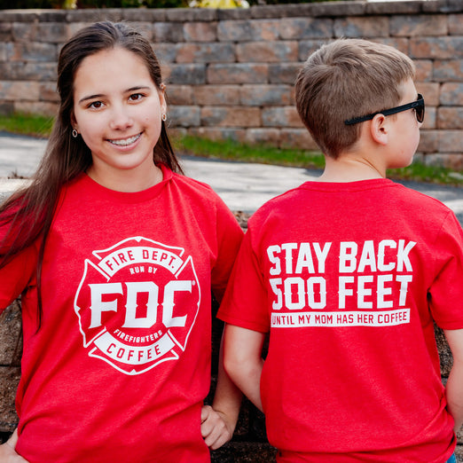 A girl and a boy standing next to with other in red shirts that have the FDC maltese cross logo on the front and the back reads, ”Stay back 500 feet until my mom has her coffee”