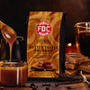 FDC Butter Toffee Bourbon coffee bag with a cup of coffee and caramel sauce on a wooden surface.