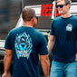 Two men wearing blue t-shirts with logos and text, standing in front of a building with a sign.