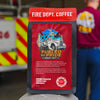 Fire Department Coffee package with a fire truck illustration and text, set against a blurred background of a fire truck and person.