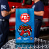 A blue bag of FDC Fire Department Rapid Response coffee stands on a diamond plate table, with blurred firefighters and a red fire truck in the background.