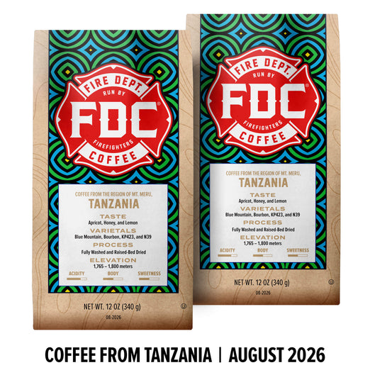 Two bags of FDC Fire Department Coffee from Tanzania displayed side by side on a white background with a black text banner at the bottom reading ’COFFEE FROM TANZANIA | AUGUST 2026’. The bags feature a light wood grain texture with a colorful geometric pattern in blue, green, and yellow, and a prominent red and white FDC logo.
