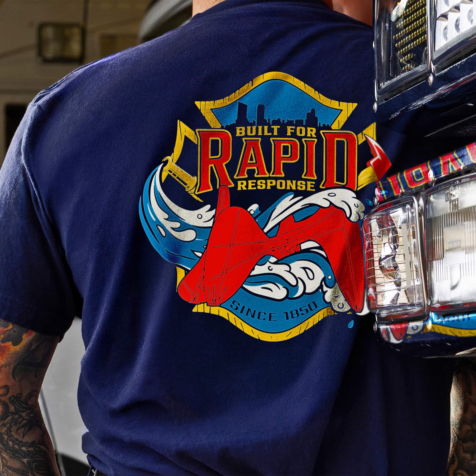 A navy blue short-sleeve t-shirt shown from the back, featuring a large graphic print with a firefighter helmet, red helmet, and the words 'Built for Rapid Response' on the back, and 'FDG' on the sleeve. A black banner at the bottom reads 'GIVES BACK TO SUPPORT' with a smaller version of the fire department emblem in the lower right corner.