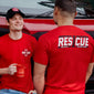 Two men wearing red ’Rescue’ t-shirts with a coffee cup in front of a vehicle.