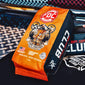 A bag of FCX Combat Coffee Club coffee lies on a black surface with a dark metal mesh background. The packaging is bright orange on the left with a cartoon tiger mascot and the words ’Fort Together Battle As One,’ and black on the right with large white ’CLUB’ lettering and a small red ’FCX’ logo.