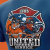 Close-up of a blue T-shirt showing a large firefighter-themed graphic with a tiger and a rooster in turnout gear, flames, water, and the text 'SINCE 1905' and 'UNITED THROUGH SERVICE'.