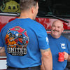 Two men in blue firefighter-themed t-shirts standing in front of a red fire truck. The man on the left has his back to the camera, showing a large graphic on the back with the text 'UNITED THROUGH SERVICE' and 'FIRE DEPT.