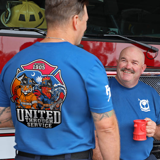 Two men in blue firefighter-themed t-shirts standing in front of a red fire truck. The man on the left has his back to the camera, showing a large graphic on the back with the text ’UNITED THROUGH SERVICE’ and ’FIRE DEPT.