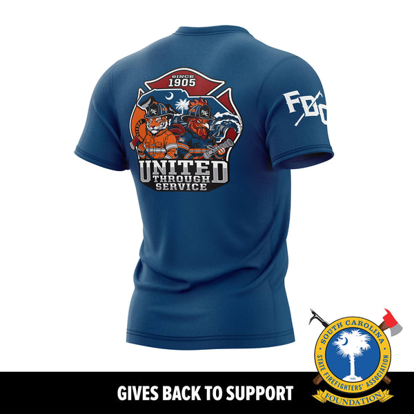 A blue short-sleeve t-shirt shown from the back, featuring a large graphic with firefighters and the text 'United Through Service 1905,' plus a white 'FOG' print on one sleeve. A black banner at the bottom reads 'GIVES BACK TO SUPPORT' with a South Carolina Fire Fighters Foundation logo.