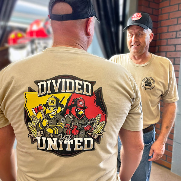 A tan short-sleeve T-shirt shown from the back on a white background, with a large firefighter-themed graphic reading 'DIVIDED BUT UNITED' and a fire department badge on the back, plus partial sleeve text. A black banner at the bottom reads 'GIVES BACK TO SUPPORT' and includes a fire department emblem.