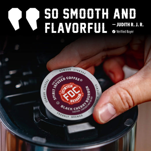 A hand inserts a single K-Cup coffee pod into a black coffee maker. The pod is labeled FDC Freshly Brewed Coffee, Spirit Infused Coffee, and Black Cherry Burger.