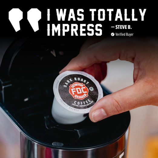 A hand inserts a single FDC Coffee K-Cup pod into a black coffee maker. The pod label reads ’Dark Roast FDC Coffee’ with a red and white fire department badge.