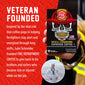 A hand holds a box of Fire Department Coffee ’Skull-Crushing Espresso Coffee’ against a red textured background with white text reading ’VETERAN FOUNDED’ and a short descriptive blurb. The box features a skull wearing a firefighter helmet, and a small circular inset photo shows a man in a white sailor uniform.