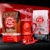 The Coffee Lovers Valentine's Day Bundle, containing a bag of Donut Shop Coffee, a bag of Strawberries and Cream Rum Infused Coffee, a red EKG Sticker, and a red Fire Hydrant mug set on the front bumper of a red fire truck.