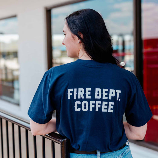 A lifestyle image of someone wearing a Crew T Shirt. The back of the navy shirt says ”Fire Dept. Coffee” in white text.