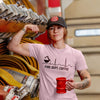 A woman wearing a pink Fire Dept. Coffee EKG T-shirt and black FDC hat, flexing beside coiled fire hoses while holding a red fire hydrant mug. The shirt design features a black coffee pot pouring into a heartbeat line with “Fire Dept. Coffee” text underneath.