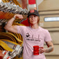 A woman wearing a pink Fire Dept. Coffee EKG T-shirt and black FDC hat, flexing beside coiled fire hoses while holding a red fire hydrant mug. The shirt design features a black coffee pot pouring into a heartbeat line with “Fire Dept. Coffee” text underneath.