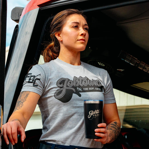 A lifestyle image of someone wearing the Jobtown Shirt. This grey shirt features ”Jobtown” on the front in a script font and text underneath that reads, ”Fire Dept. Coffee”