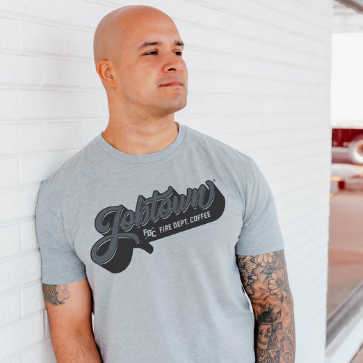 A lifestyle image of someone wearing the Jobtown Shirt. This grey shirt features ”Jobtown” on the front in a script font and text underneath that reads, ”Fire Dept. Coffee”