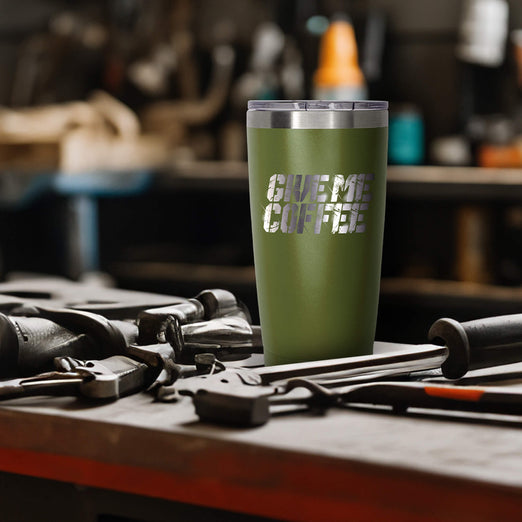 A green tumbler that says GIVE ME COFFEE on the front. The tumbler is sitting on a table with tools around it.