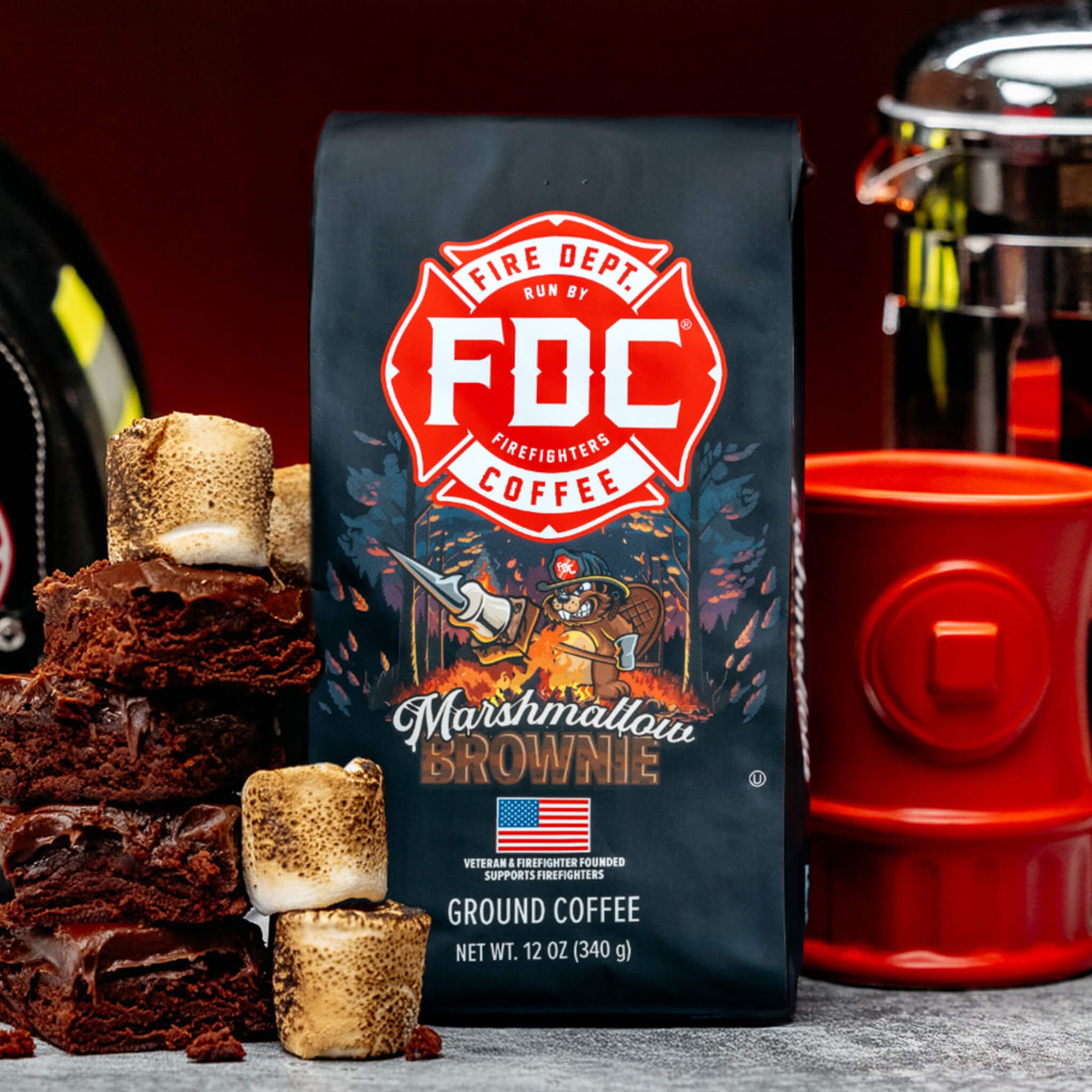 A bag of Fire Department Coffee's 12 oz Marshmallow Brownie Coffee with a red section accross the top that says "Fire Dept. Coffee". The art on the bag features a beaver holding a pike pole with a marshmallow and brownie on it over a campfire. The title reads, "Marshmallow Brownie" and there is a veteran owned badge with an American flag in the bottom right corner.