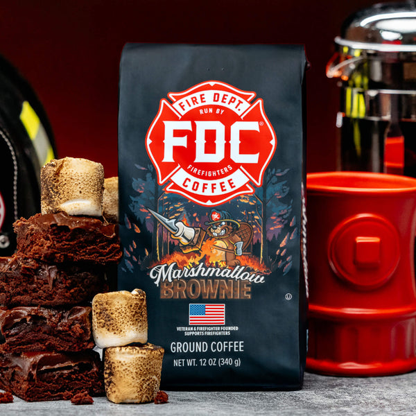 A bag of Fire Department Coffee's 12 oz Marshmallow Brownie Coffee with a red section accross the top that says "Fire Dept. Coffee". The art on the bag features a beaver holding a pike pole with a marshmallow and brownie on it over a campfire. The title reads, "Marshmallow Brownie" and there is a veteran owned badge with an American flag in the bottom right corner.