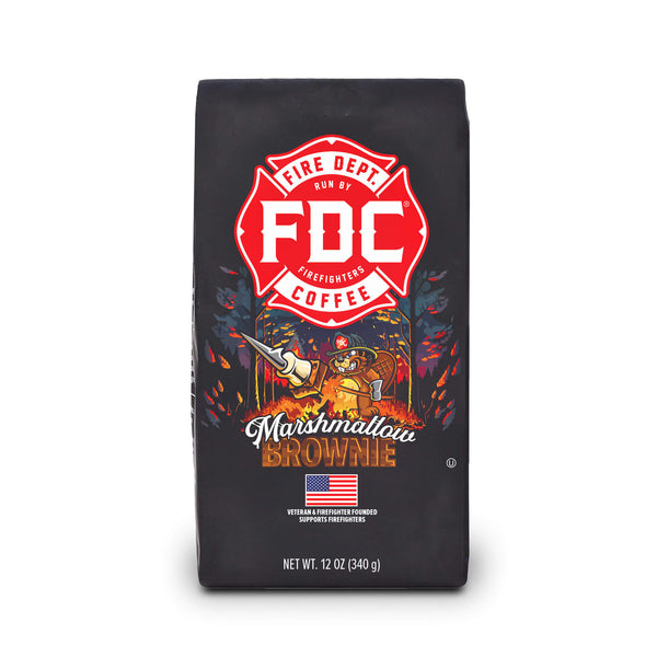 A bag of Fire Department Coffee's 12 oz Marshmallow Brownie Coffee with a red section accross the top that says "Fire Dept. Coffee". The art on the bag features a beaver holding a pike pole with a marshmallow and brownie on it over a campfire. The title reads, "Marshmallow Brownie" and there is a veteran owned badge with an American flag in the bottom right corner.