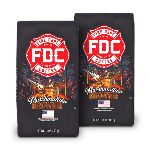 Two bags of Fire Department Coffee’s 12 oz Marshmallow Brownie Coffee with a red section accross the top that says ”Fire Dept. Coffee”. The art on the bag features a beaver holding a pike pole with a marshmallow and brownie on it over a campfire. The title reads, ”Marshmallow Brownie” and there is a veteran owned badge with an American flag in the bottom right corner.