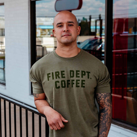 A lifestyle image of someone wearing the FDC Military Green Shirt. The front of the shirt says ”Fire Dept. Coffee” in dark green lettering.