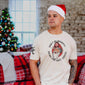 Someone wearing a Don’t Be Naughty Shirt, wearing a Santa hat with a Christmas tree behind them