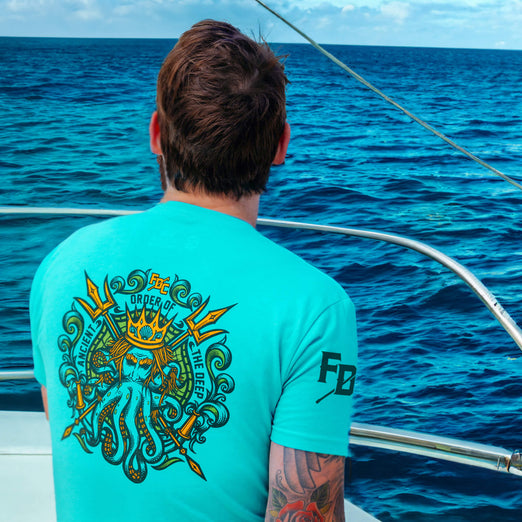 A lifestyle image of someone wearing the Shellback Shirt. The back of the shirt features imagery of King Neptune and says ”Ancient Order of the Deep” with the FDC pike pole logo above in yellow.
