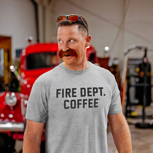 Man wearing a gray ’Fire Dept. Coffee’ text t-shirt in a fire station with a vintage red truck in the background.