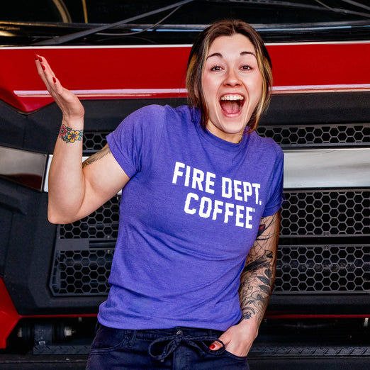 A woman wearing a purple t-shirt with white ’FIRE DEPT. COFFEE’ text in front of a red and black fire truck.