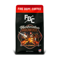 A bag of Fire Department Coffee’s 12 oz Marshmallow Brownie Coffee with a red section accross the top that says ”Fire Dept. Coffee”. The art on the bag features a beaver holding a pike pole with a marshmallow and brownie on it over a campfire. The title reads, ”Marshmallow Brownie” and there is a veteran owned badge with an American flag in the bottom right corner.