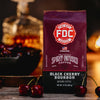 A bag of Black Cherry Bourbon Infused Coffee sitting on a table surrounded by cherries with a decanter of bourbon in the background