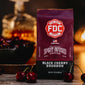 A bag of Black Cherry Bourbon Infused Coffee sitting on a table surrounded by cherries with a decanter of bourbon in the background