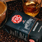 A bag of Bourbon Infused Coffee surrounded by coffee beans and a glass of bourbon