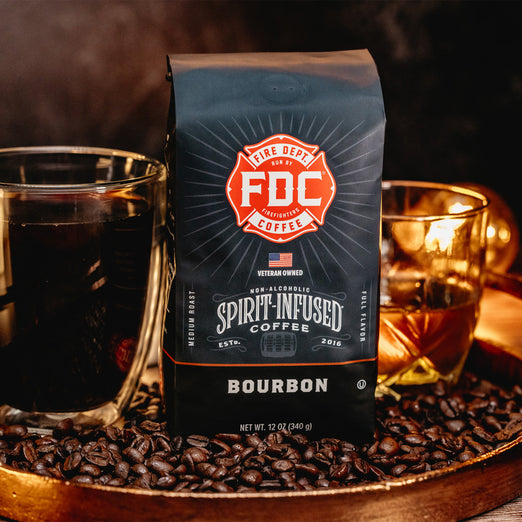 A bag of Bourbon Infused Coffee sitting among coffee beans with a glass of coffee next to it and a glass of bourbon behind it