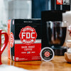 A box of Dark Roast Coffee Pods sitting on a table with a cup of coffee behind it.