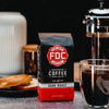 An image of FDC's Dark Roast on a table surrounded by coffee brewing items.