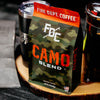 A photo of a bag of Fire Dept Coffee's Camo Blend on a tree stump surrounded by various camping tools.