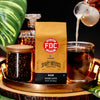 Fire Dept. Coffee's 12 ounce Rum Infused Coffee on a tray surrounded by a brewed cup of coffee with milk and tropical plant leaves.