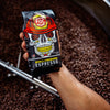 A 12oz bag of Skull Crushing Espresso being held over a roaster full of roasted coffee beans.